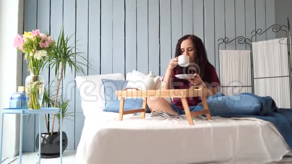 早上穿着睡衣躺在床上的年轻迷人的女人漂亮的女人坐在家里的大床上喝咖啡视频的预览图