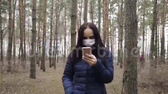 年轻女子戴着医用口罩在森林里散步在手机上了解疫情的消息成年女性视频的预览图