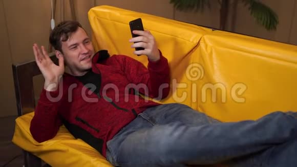 穿休闲服装的年轻人躺在黄色沙发上成年男性在手机中休息和翻阅各种新闻视频的预览图