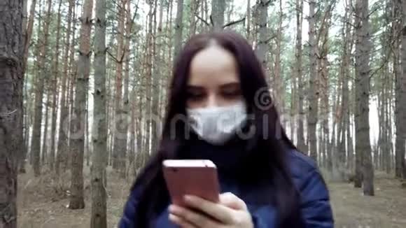 年轻女子戴着医用口罩在森林里散步在手机上了解疫情的消息成年女性视频的预览图