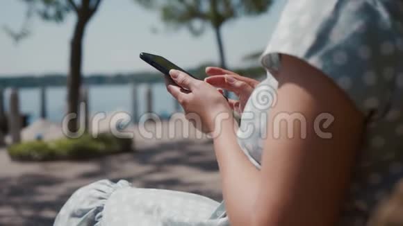 穿着蓝色波尔卡多礼服的女人坐在公园长凳上用智能手机关门视频的预览图