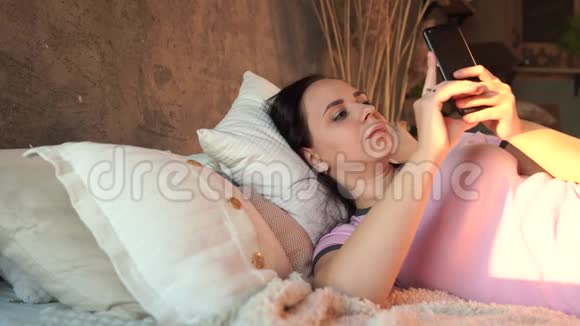 快乐的年轻女人在床上使用智能手机从上面看快乐的年轻女性一边看镜头一边浏览视频的预览图