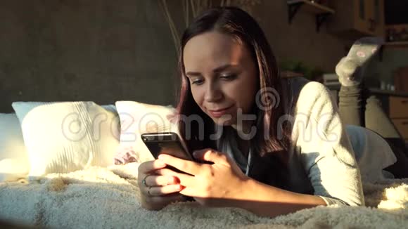 快乐的年轻女人在床上使用智能手机从上面看快乐的年轻女性一边看镜头一边浏览视频的预览图