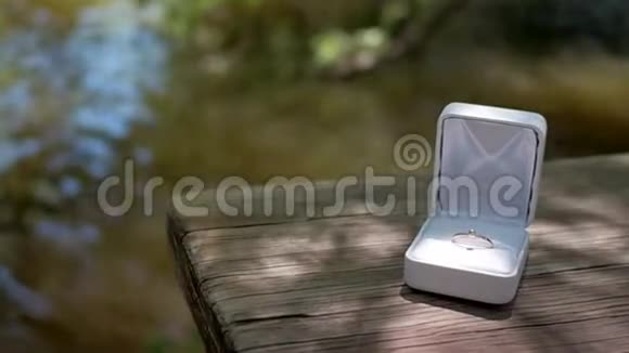 白色盒子里的金色订婚戒指坐在河边的木桌上视频的预览图