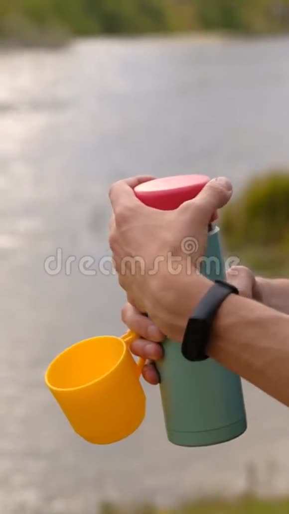 男性旅行者打开一个热水瓶把茶倒入河底的黄色塑料杯中视频的预览图