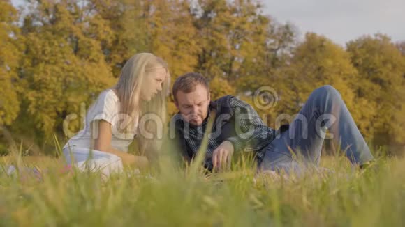 英俊的白种人男人躺在秋草上给一个年轻的金发女孩看书父女穿着视频的预览图