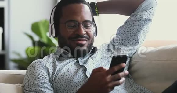 戴着耳机听沙发上的移动音乐的快乐非洲男人视频的预览图