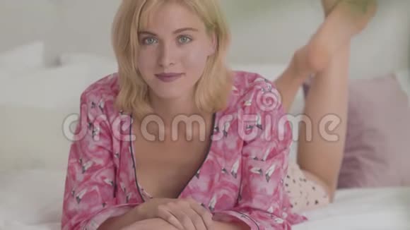 年轻的金发白种人女孩灰色的眼睛穿着粉红色的睡衣躺在白色柔软的床上看着相机视频的预览图