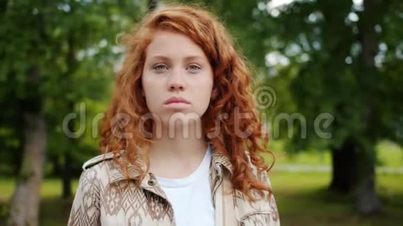 严肃的少年红发女孩正站在公园的室外看着摄像机视频的预览图