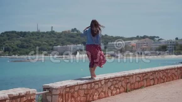 戴着太阳镜穿着浪漫的红色连衣裙和牛仔夹克的快乐的年轻女子走在堤岸的石墙上视频的预览图