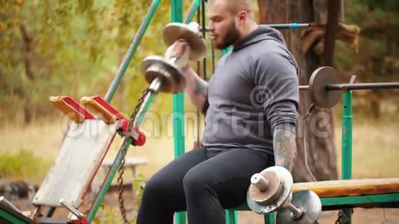 一名纹身男健美运动员将哑铃拉在双手上哑铃被拴在长凳上训练视频的预览图