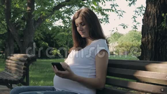 怀孕妇女在夏日坐在公园长椅上使用智能手机应用程序视频的预览图
