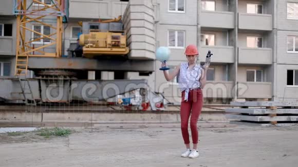 戴着钻和地球仪的建筑头盔的年轻女子在建筑中的房子和起重机的背景下跳舞视频的预览图