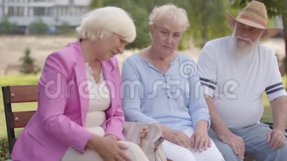 三个成熟的白种人朋友坐在夏季公园的长凳上一头金发的老女人抱怨视频的预览图