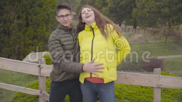 戴眼镜的白种人少年用黄色外套抚摸他微笑的怀孕母亲的腹部快乐的母亲视频的预览图