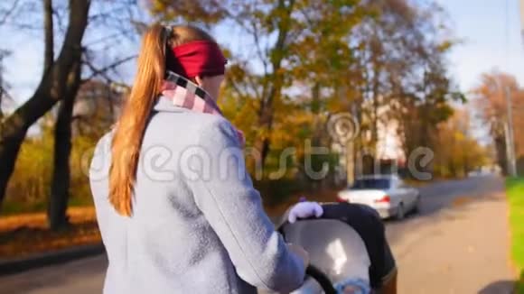 快乐的年轻妈妈和宝宝一起走在公园秋天的婴儿车里微笑着走进镜头产妇概念视频的预览图