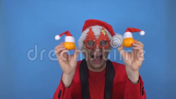 一个穿着圣诞老人服装的白种人年轻人手里拿着橘子跳舞看着镜头蓝色视频的预览图