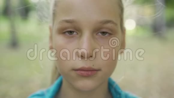 一个年轻的金发女孩看着相机的特写肖像美丽的白种人少年站在阳光明媚的公园里视频的预览图