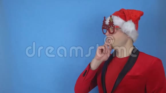 一个戴着圣诞眼镜和圣诞老人服装的年轻人在蓝色背景下思考和看着摄像机视频的预览图