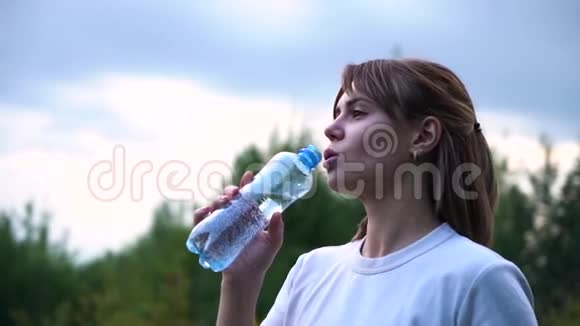 年轻漂亮的运动女孩穿着一件白色T恤很累喝着透明瓶子里的水慢动作视频的预览图