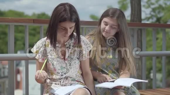 两个漂亮的女学生坐在户外一起讨论家庭作业写在笔记本上夏季视频的预览图
