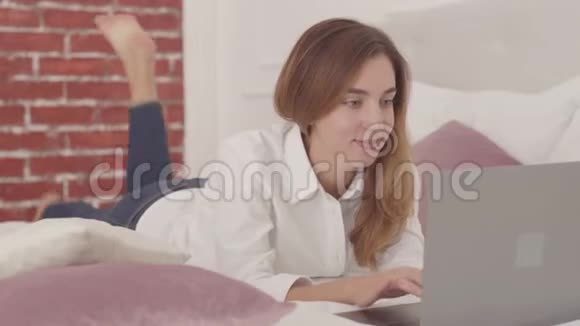带着笔记本电脑和打字躺在床上的白人女孩微笑漂亮的女学生在家随意学习视频的预览图