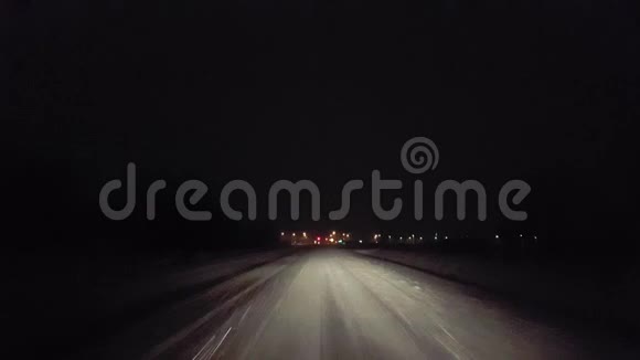 夜间在乡村道路上下雪时驶近城市交通灯POV向城市移动的驱动视角视频的预览图