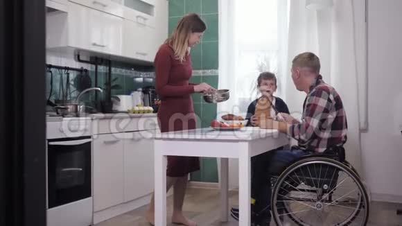 家庭晚餐坐在轮椅上的快乐父亲和微笑的妻子和儿子在厨房准备食物视频的预览图