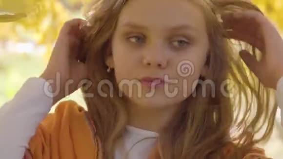 一个可爱的金发白种人女孩戴着芥末围巾站在秋天公园的黄叶后面儿童视频的预览图