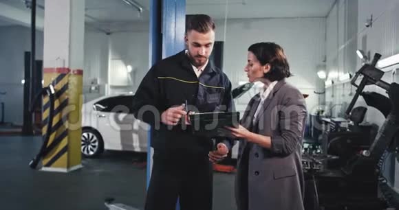在一个大型的汽车服务中心一个有魅力的机械师穿着制服和一个女商人在一些文件上签名然后他们就来了视频的预览图