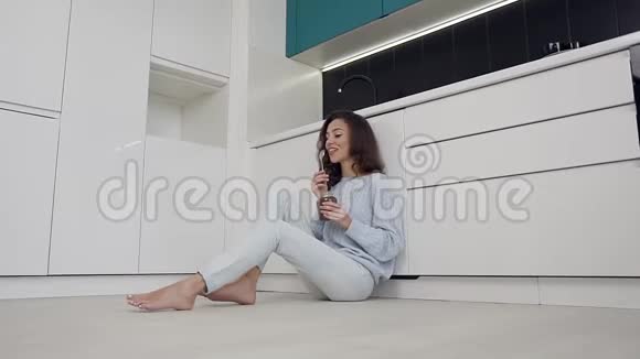 可爱的现代微笑女人穿着时髦的衣服坐在厨房的地板上吃着罐子里的巧克力奶油视频的预览图