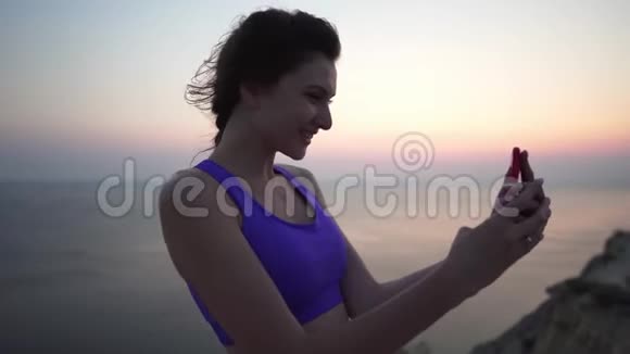 美丽的年轻运动女孩穿着紫色上衣在日落或日出的海洋背景下自拍A视频的预览图