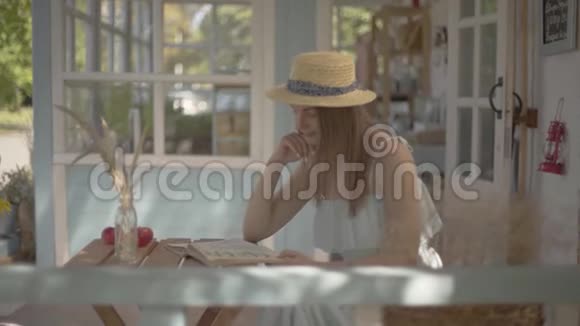 可爱的年轻农村女孩戴着草帽和白色连衣裙坐在户外的小木桌上看书农村视频的预览图