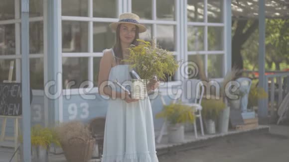 可爱的年轻女子戴着草帽穿着白色连衣裙微笑着看着镜头嗅着前面的野花视频的预览图