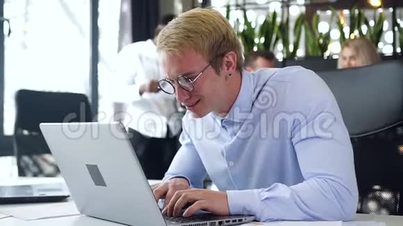 漂亮的金发年轻商人戴着眼镜坐在办公室里在电脑上完成了他的工作视频的预览图