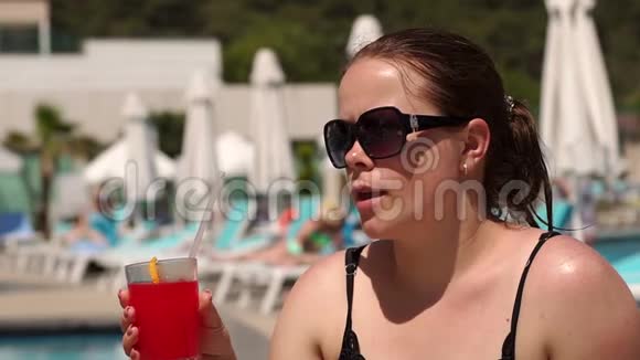 一个戴墨镜的女孩坐在游泳池旁的休息室里喝鸡尾酒视频的预览图