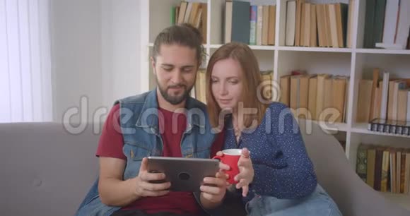 一对年轻夫妇在玩平板电脑游戏在家里被吸收并感兴趣视频的预览图