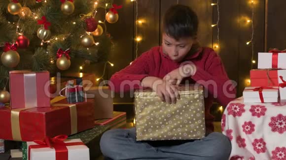 男孩坐在室内靠近装饰的圣诞树灯光和打开礼物圣诞快乐和节日快乐视频的预览图