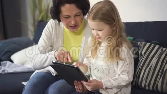 成年妇女坐在沙发上教小女孩使用新的数字技术视频的预览图