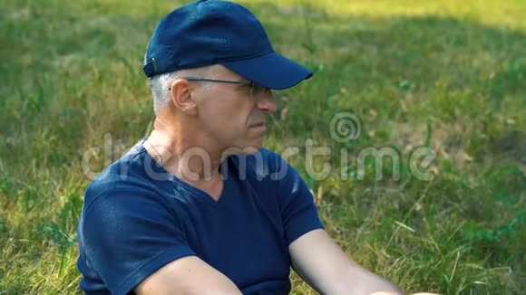 一个夏天一个身材苗条头发严肃穿着蓝色t恤帽子和眼镜的人坐在森林里的绿草上视频的预览图