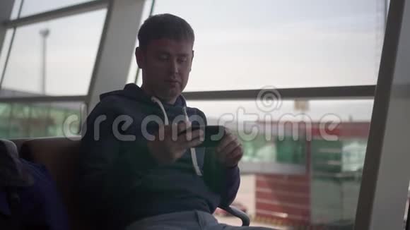 美国男子坐在机场坐在帕诺曼的窗户椅子上看手机上的应用程序视频的预览图
