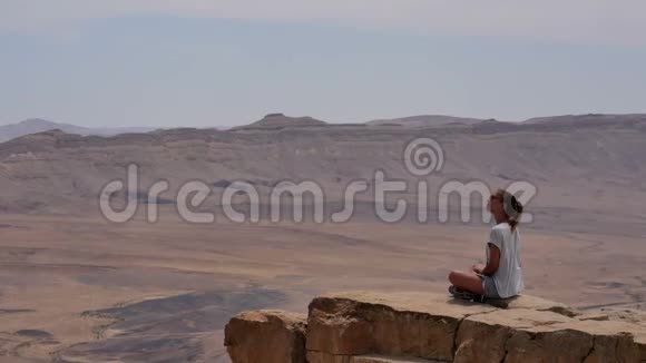 戴墨镜的年轻女子坐悬崖边环顾沙漠视频的预览图