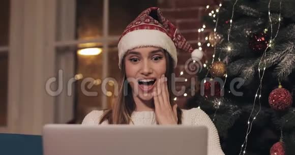 美丽的金发白种人女孩戴着桑塔帽坐在圣诞树附近用她的笔记本电脑聊天视频的预览图