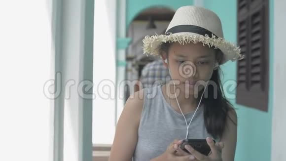 亚洲可爱的女孩穿着休闲装戴着草帽用手机和耳机悠闲地走在街边视频的预览图