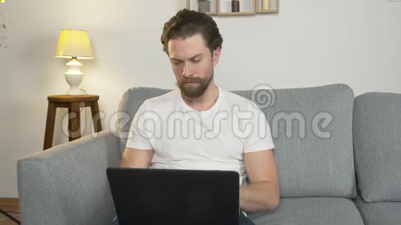 一个年轻人独自坐在家里的沙发上坐在笔记本电脑上上网冲浪因为视频的预览图
