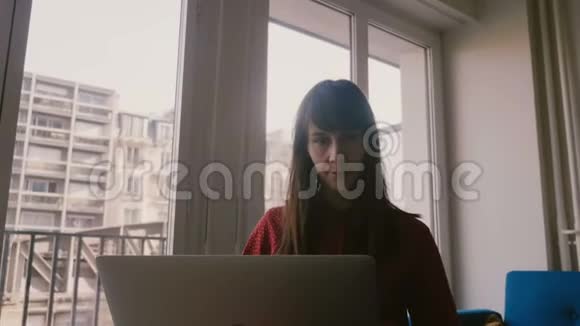 在巴黎出差时漂亮的专注年轻女商人在公寓窗口附近使用笔记本电脑和智能手机视频的预览图