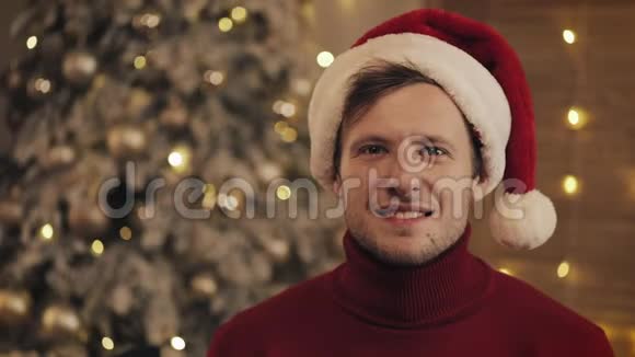 戴圣诞帽戴冬红毛衣笑容可掬的年轻人坐在圣诞树上的画像视频的预览图