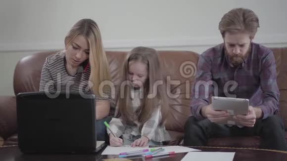 可爱的小女孩坐在母亲和父亲之间用彩色标记画画年轻女子在她的笔记本电脑上打字视频的预览图