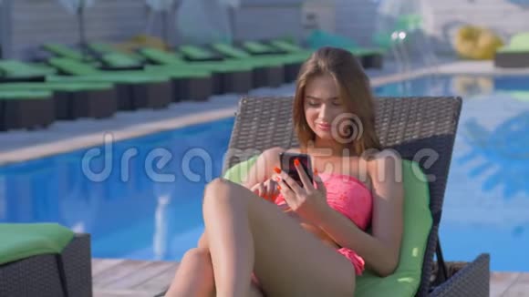 度假村度假漂亮的年轻女孩穿着泳衣在太阳休息室的蓝色游泳池附近休息看着一个电话视频的预览图
