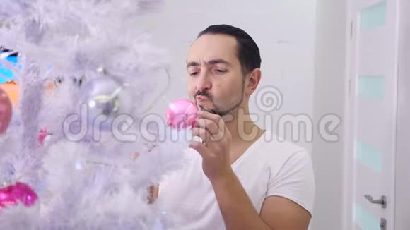 在皮贾马的男人装饰圣诞树看着他的倒影在粉红色的玻璃泡视频的预览图